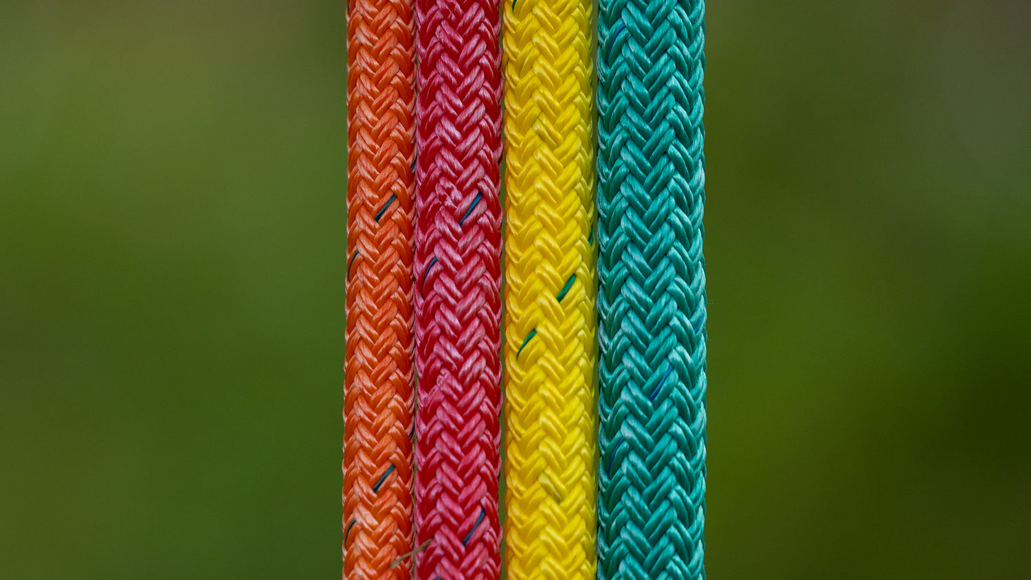 Four close-up vertical strands of braided rope or cord in orange, red, yellow, and teal, set against a blurred green background.