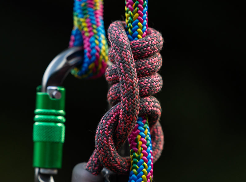 A close-up of a knotted multicolor climbing rope with a green carabiner and metal hardware against a dark blurred background.
