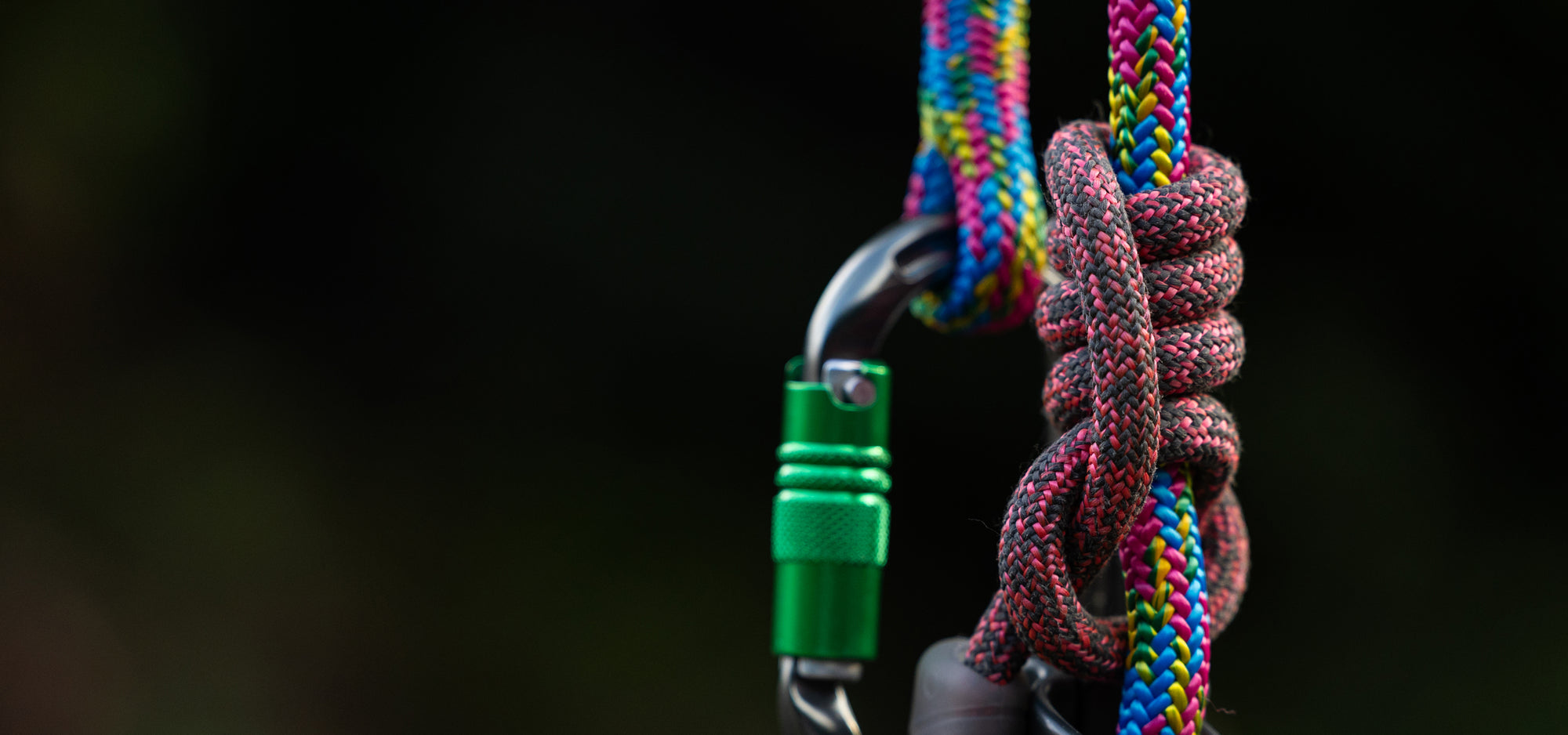 Close-up of a multicolored climbing rope tied in a knot with a metal carabiner and green locking device against a dark blurred background.