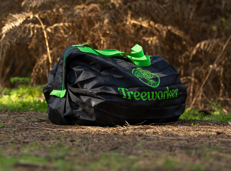 A black duffel bag with bright green handles and the 'Treeworker' logo printed on the front, sitting on a dirt ground with ferns in the background.