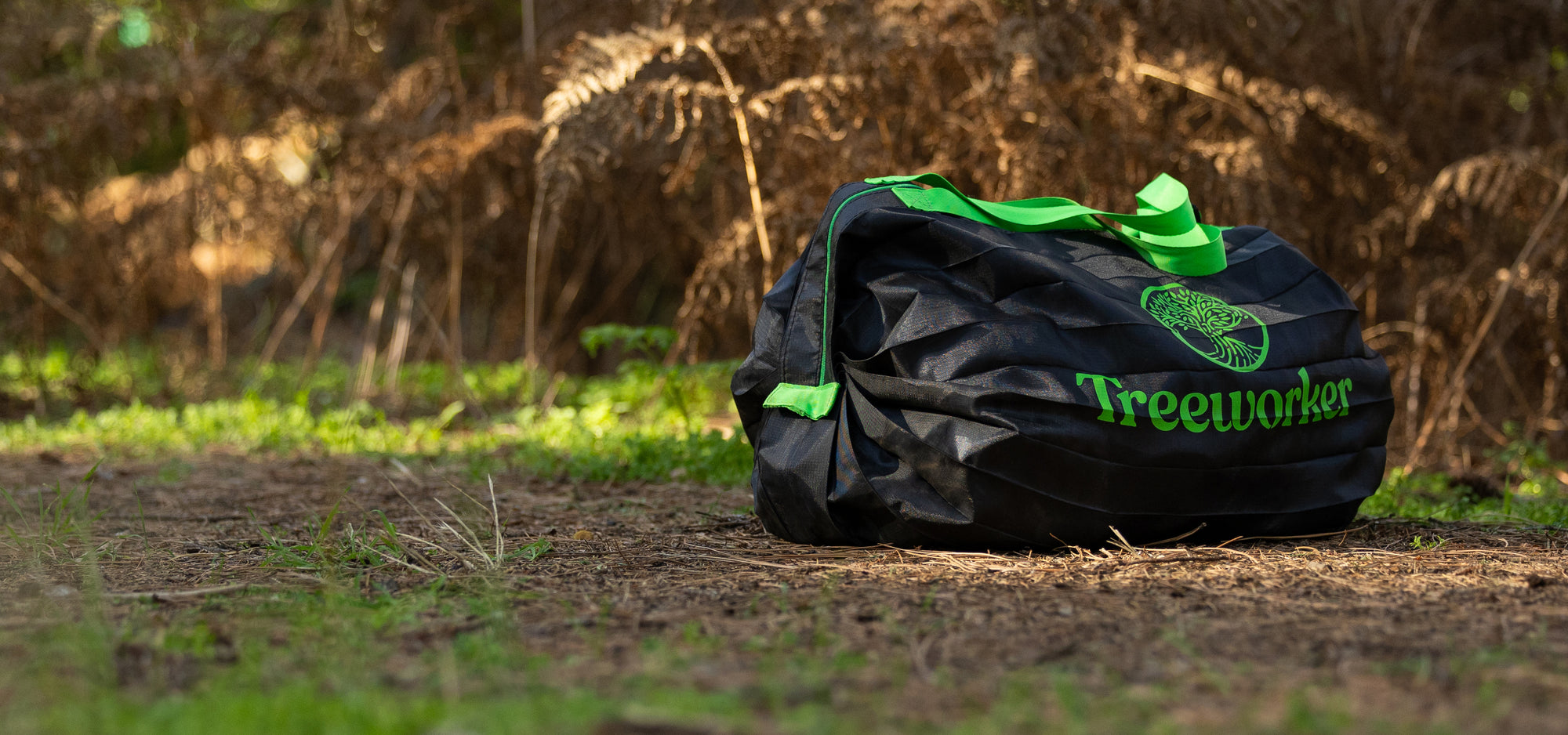 A black duffel bag with bright green handles and the 'Treeworker' logo printed on the side, resting on a dirt path with a blurred forest background.