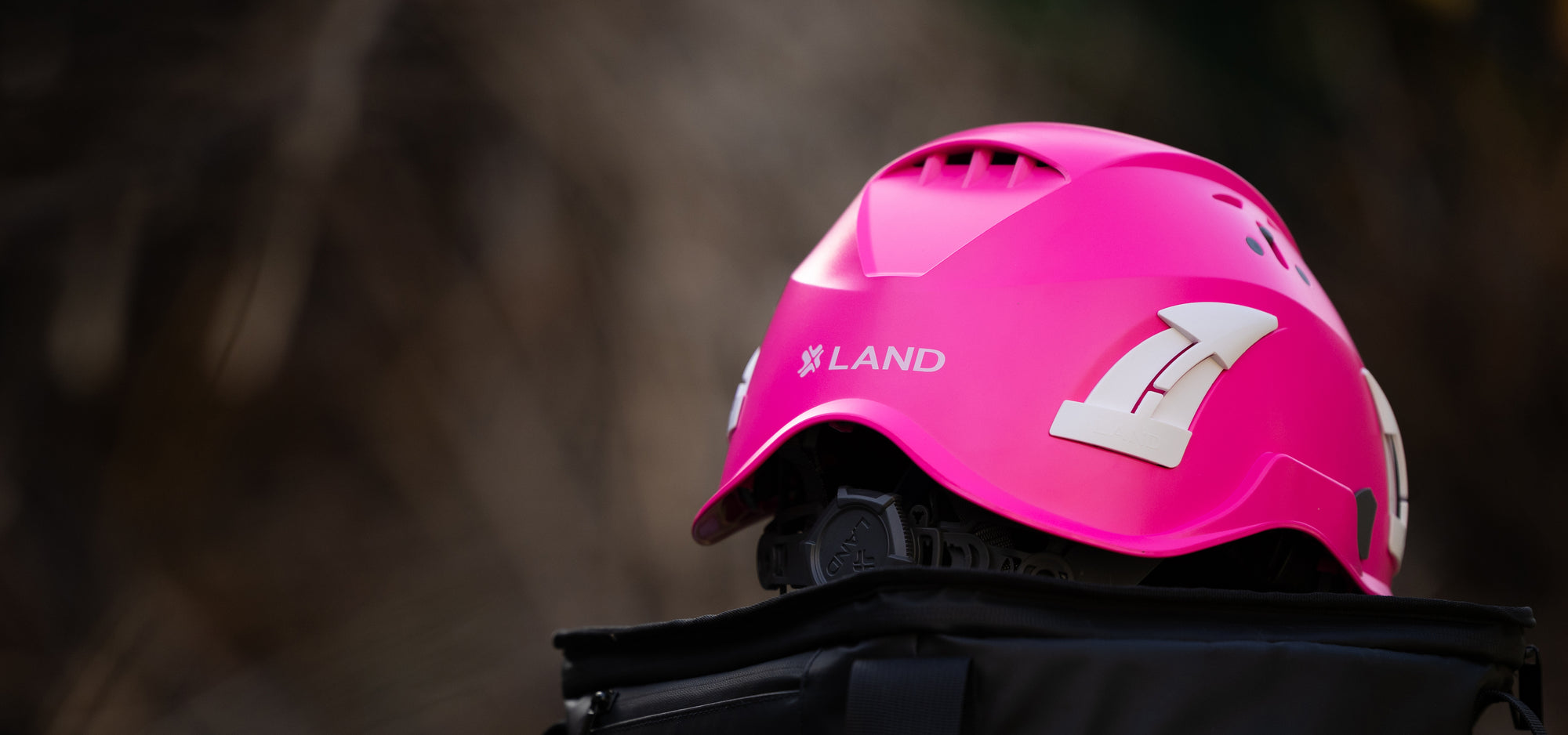 A bright pink climbing helmet with white graphics and the brand name LAND, resting on a black bag against a blurred outdoor background.