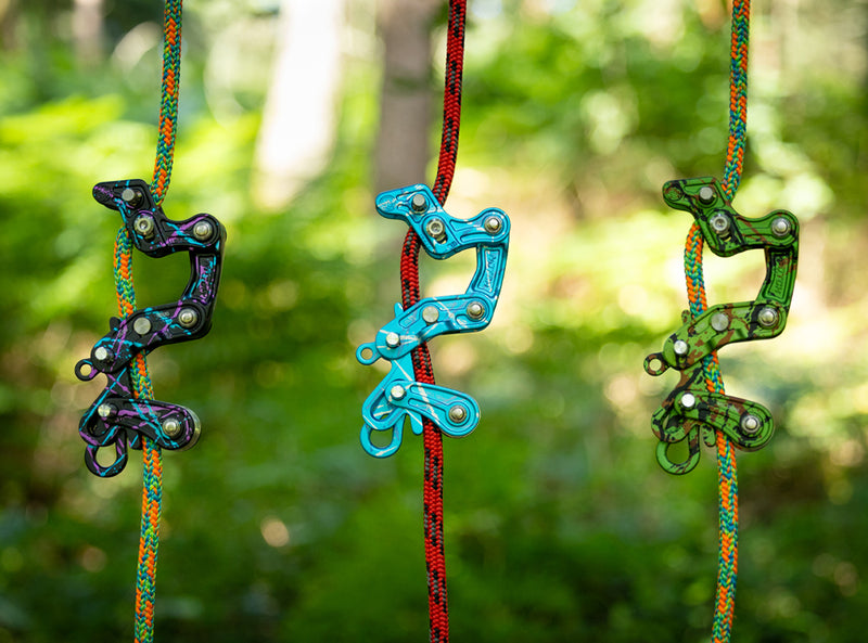 Three colorful climbing or rappelling hardware pieces hanging from braided ropes in a blurred green outdoor setting.