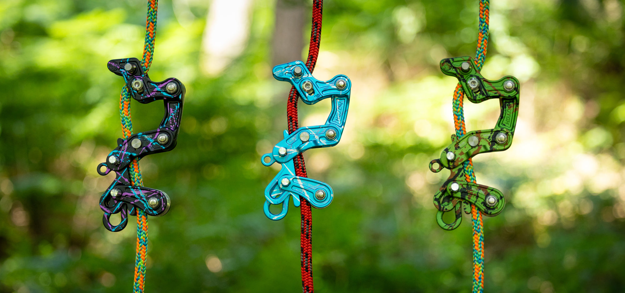 Three colorful climbing or rappelling quickdraws hanging vertically against a blurred green forest background. The left draw is black with multicolor flecks, the center draw is bright blue, and the right draw is green with multicolor flecks.